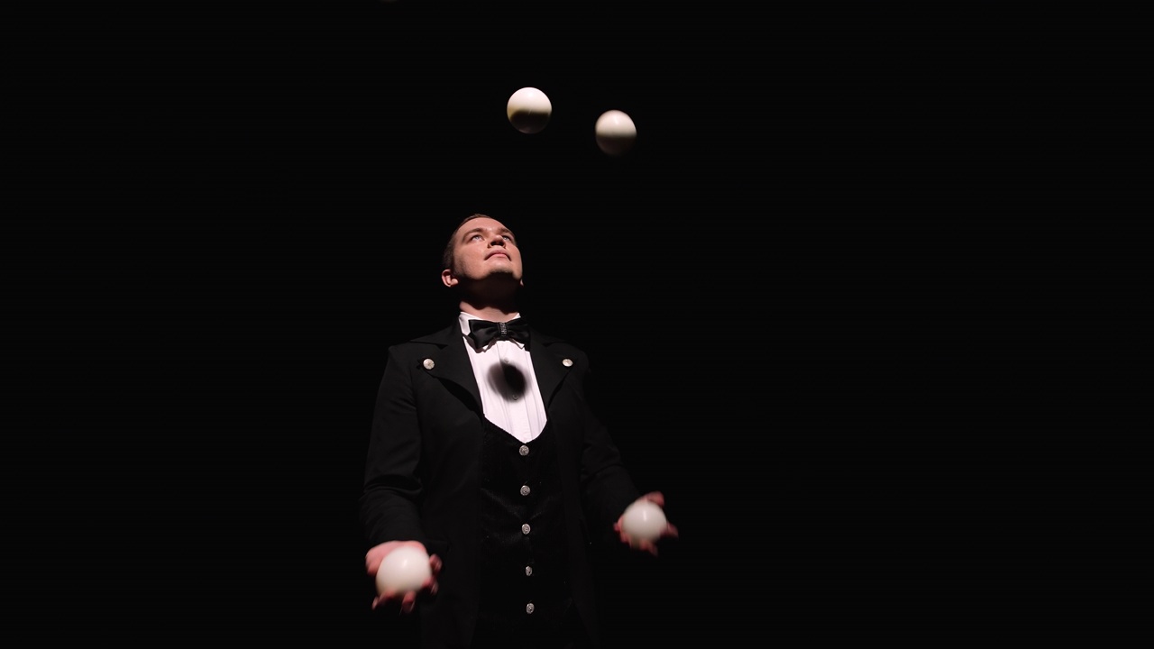 轨道照片上一个穿黑西装的人在职业抛白球。一个马戏团演员在灯光的照耀下，在黑暗的演播室里抛球接球。关闭了。慢动作视频下载