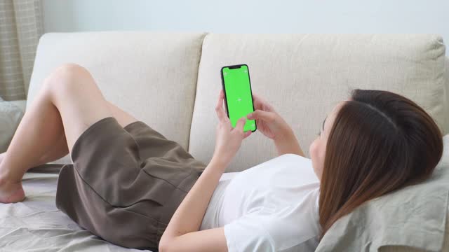 亚洲女子躺在沙发上玩绿色屏幕的智能手机视频下载