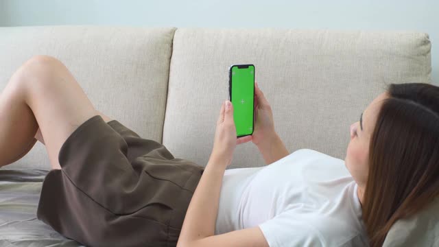亚洲女子躺在沙发上玩绿色屏幕的智能手机视频下载
