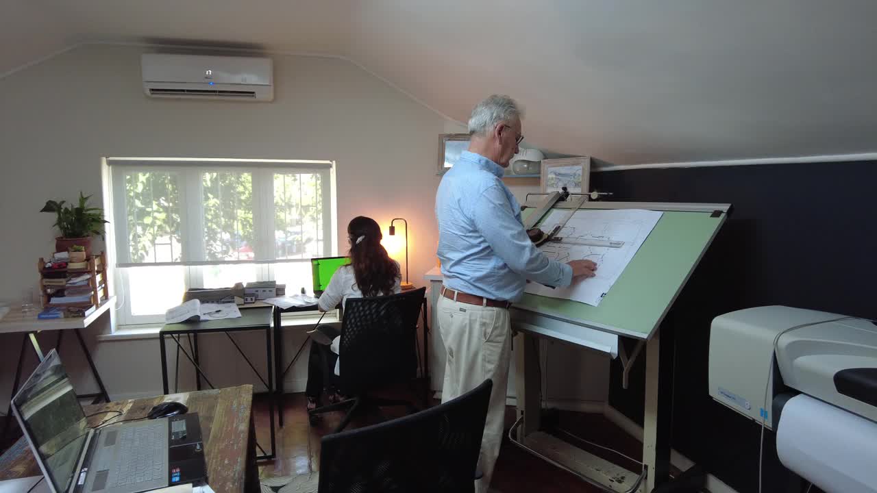侧视图拍摄的高级男建筑师工作在绘图桌移动到笔记本电脑在办公室和女建筑师后面视频下载