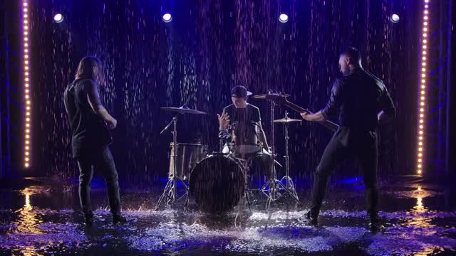 一个三人摇滚乐队在倾盆大雨中在深蓝色的灯光下表演。电吉他，贝斯和爵士鼓。现场表演在落下的雨滴。慢动作视频下载