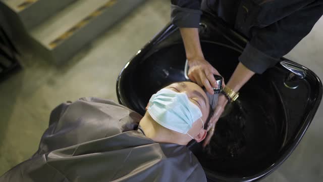 正上方戴着保护面罩的亚裔华人男子在美发沙龙里闭着眼睛躺着洗头视频素材