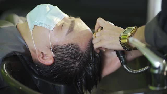 戴着保护面罩的亚裔华人男子在美发沙龙里闭着眼睛躺着洗头视频素材