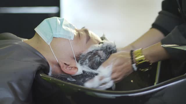 戴着保护面罩的亚裔华人男子在美发沙龙里闭着眼睛躺着洗头视频素材