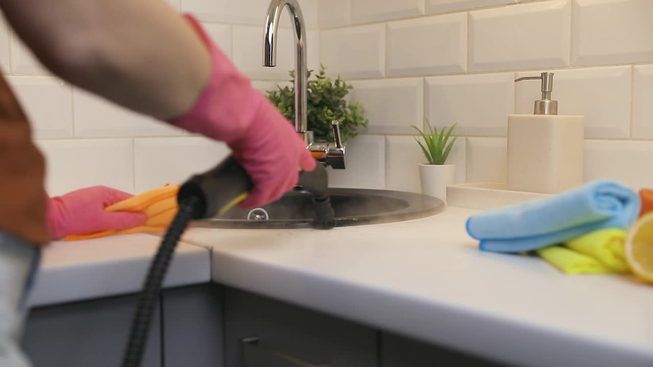 家庭主妇戴着橡胶手套，用蒸汽清洁剂清洗黑色的厨房水槽，用香瓜和海绵擦拭厨房台面。慢动作视频素材