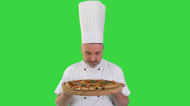 快乐的高级厨师手里拿着披萨看着绿色屏幕上的摄像头，色度键视频下载