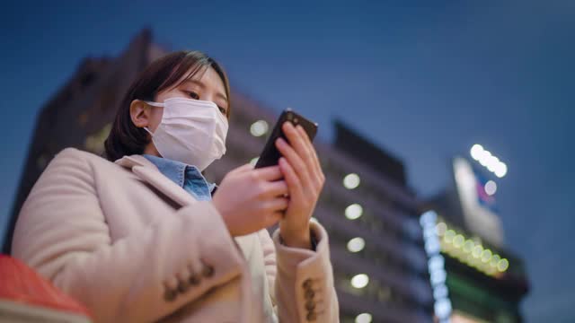 年轻女子用保护性面罩遮住脸部，晚上在城市街道上使用电话视频素材