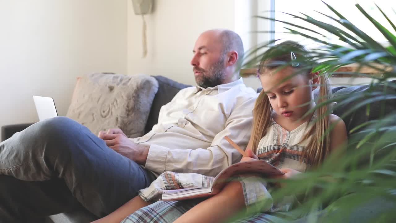父亲在用笔记本电脑，女儿坐在家里的沙发上画画视频素材