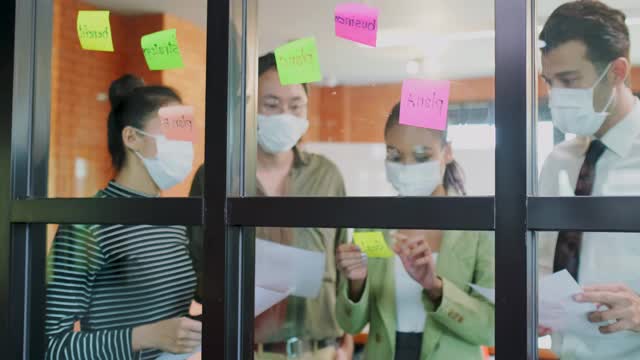 小组头脑风暴的亚洲人把便利贴贴在窗户上。团队合作的商务人士戴上口罩，可以防止新冠病毒感染，在办公室讨论和分享解决问题的想法。视频素材