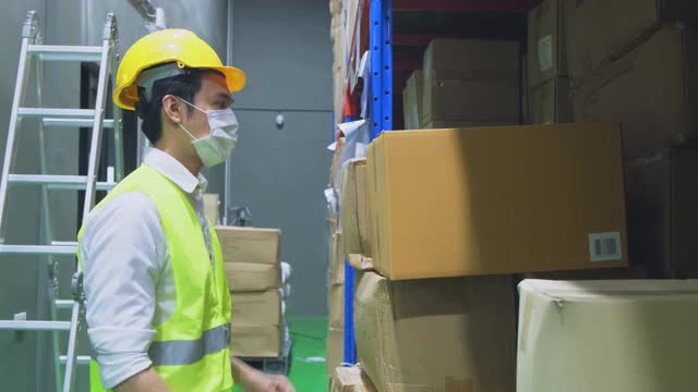 新冠肺炎流行期间，亚洲男性工人戴口罩工作。年轻人带着安全帽、背心搬运产品箱放到物流和出口工厂的货架上。视频下载