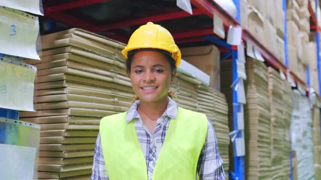 图为在新冠疫情危机期间，非裔美国女工身穿安全背心、头戴安全帽、戴着面罩站在仓库工厂内预防新冠病毒感染。视频下载