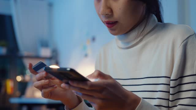 一名年轻的亚洲女性晚上坐在家里的沙发上，一边使用手机一边用信用卡进行网上交易。视频下载