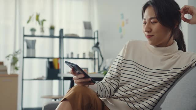 年轻的亚洲女子用手机发短信和坐在家里沙发上的朋友聊天。视频素材