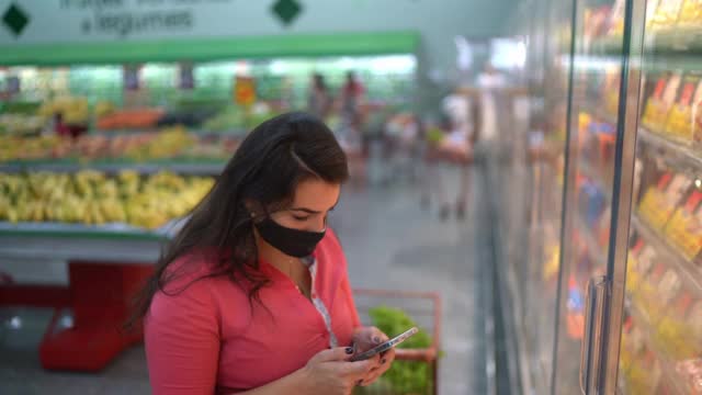 一名女子戴着口罩在超市购物时使用智能手机视频素材