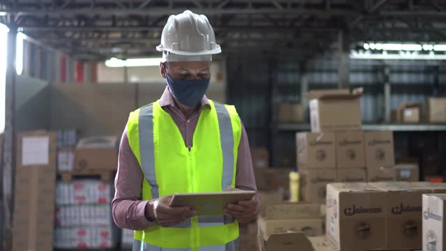 仓库工作人员戴口罩和防护服在仓库使用平板电脑检查产品视频下载