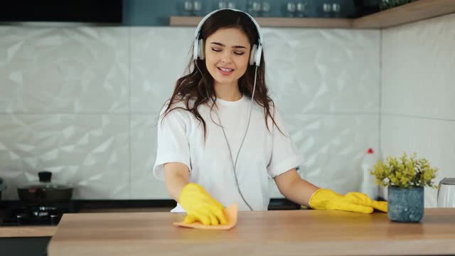 年轻妇女打扫家里。一个黑发的女模特正在厨房里清理一个灶台。女孩听音乐，打扫公寓里的灰尘视频素材