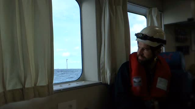 绳索访问技术人员坐在客船的客舱，从窗口看离岸农场。蓝色波浪起伏的大海和忧郁而富有戏剧性的天空。德国博库姆里夫格兰德风电场。视频素材