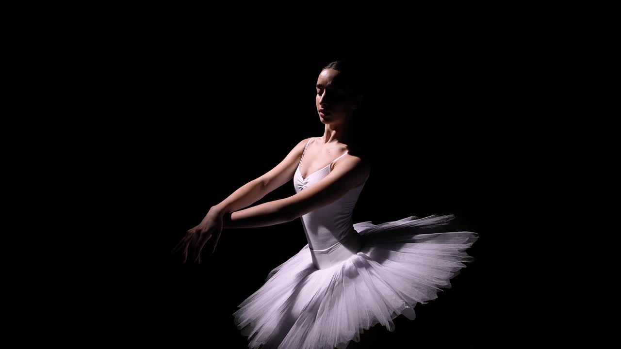 镜头围绕着穿着白色芭蕾舞裙的温柔芭蕾舞者，用她的双手表演着优雅的动作。一名年轻舞者在黑色演播室背景下翱翔的轨道镜头。关闭了。慢动作视频下载