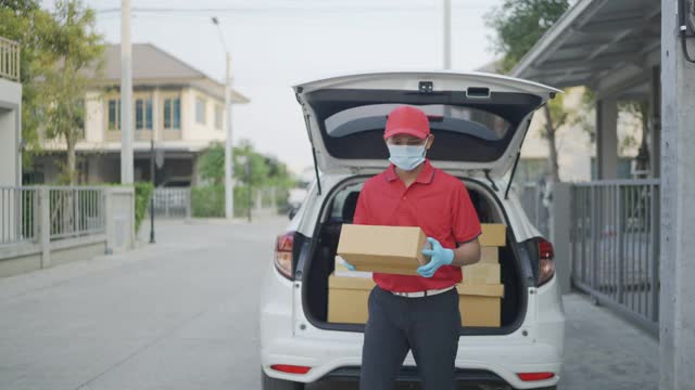 4 k慢动作视频。一名身穿红色制服的男性快递员一边送包裹一边看着摄像头。新标准的概念。视频下载