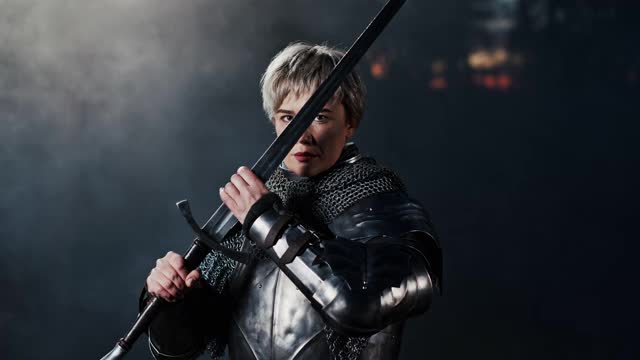 这是一个戏剧性的镜头，一个穿着中世纪金属战士盔甲的女人拿着一把剑，站在烟雾弥漫的黑暗环境中审视着剑刃视频素材