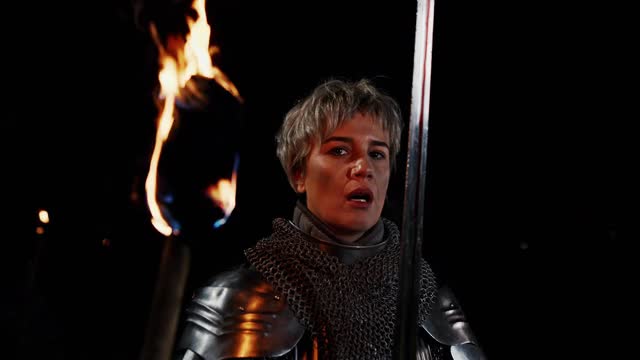 身着圣女贞德(Joan of Arc)铠甲的惊恐女子在燃烧的火炬附近呼吸，并在夜间战斗前手持宝剑离开。中世纪准备战斗的女战士视频素材