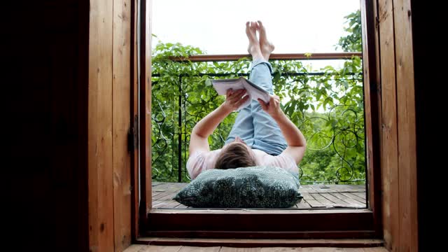 一个男人躺在阳台上，双腿翘着看书。新冠肺炎大流行期间的社交距离。在冠状病毒肆虐期间，腾出空闲时间参加最喜欢的活动视频下载