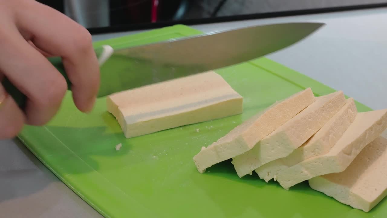 厨师右手拿着一把绿色刀柄的菜刀，在一块绿色的切菜板上慢慢地把素豆腐切成条状。特写镜头视频下载