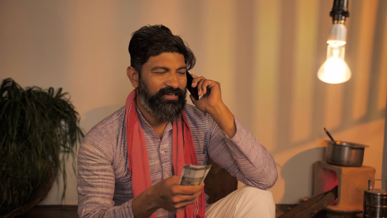 快乐的印度农民坐在家里忙着用智能手机聊天，手里拿着印度钞票视频素材
