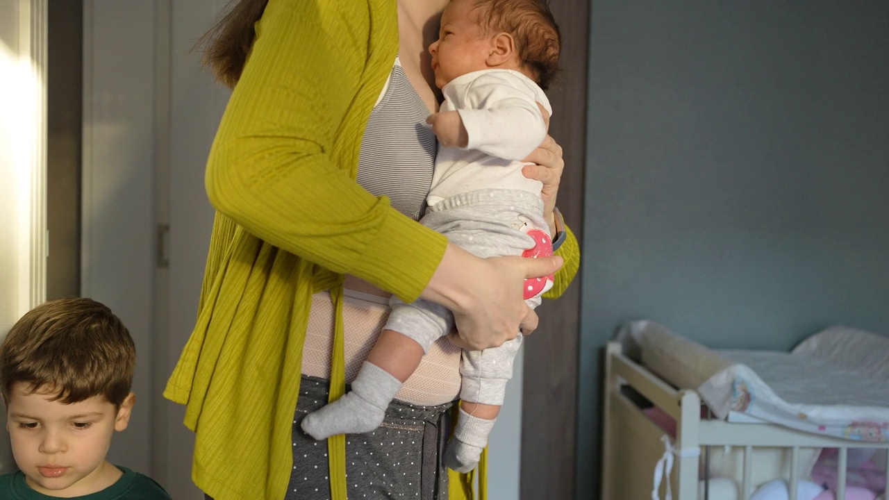 妈妈穿着产后内衣，抱着刚出生的女婴，和刚学会走路的儿子说话视频素材