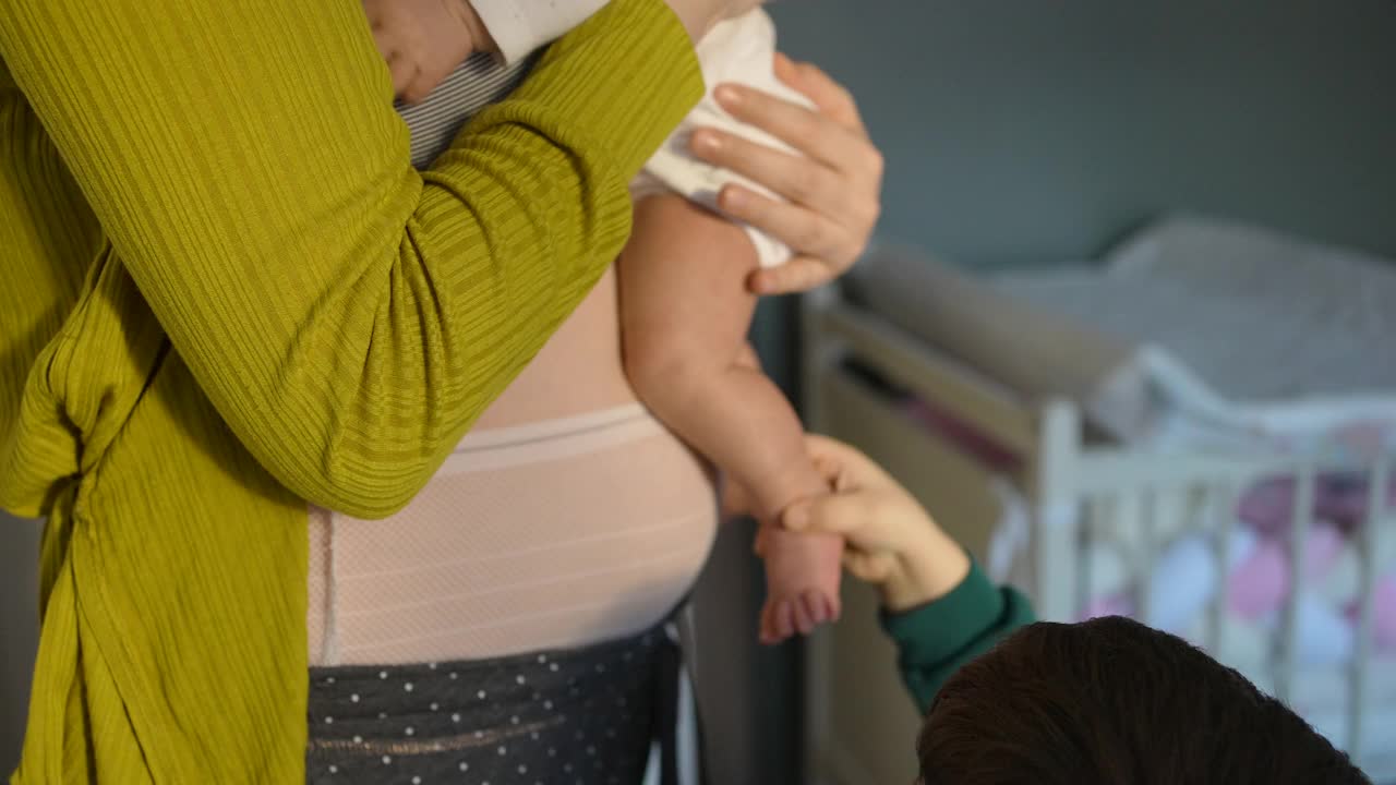 妈妈穿着产后内衣的特写，抱着刚出生的女婴，和她蹒跚学步的儿子说话视频素材