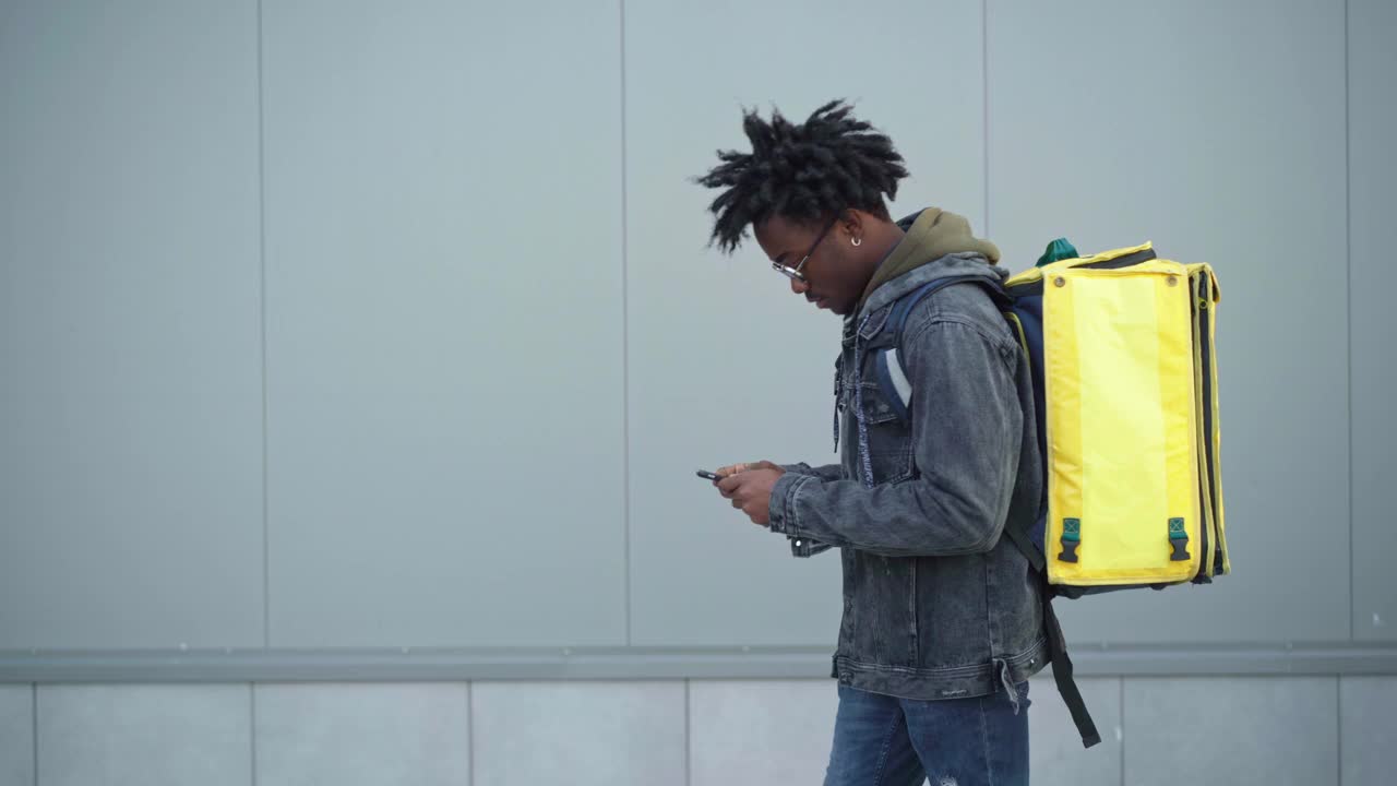 帅气的非裔美国男性快递员用智能手机挎着黄色保温包在城市中行走的侧视图。年轻人正在搜索送货地址。服务和工作理念。视频下载