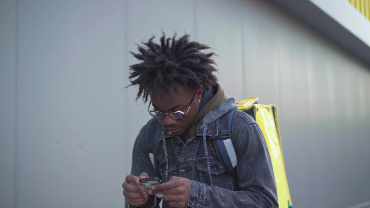 一个集中的非裔美国年轻人用智能手机查看在线地图，然后拿着黄色的绝缘食品外卖袋离开。男快递员在城市户外送信。服务的概念。视频素材