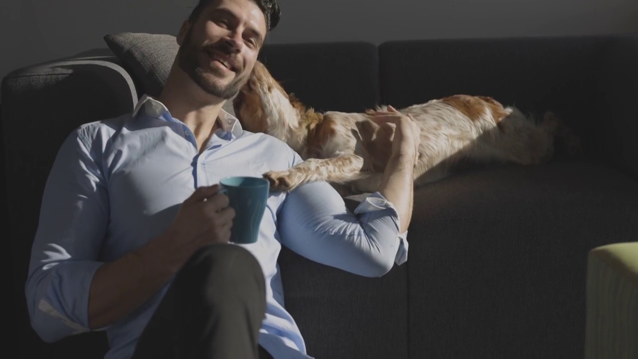 年轻的微笑男子坐在地板上，花时间与他的狗喝咖啡视频下载