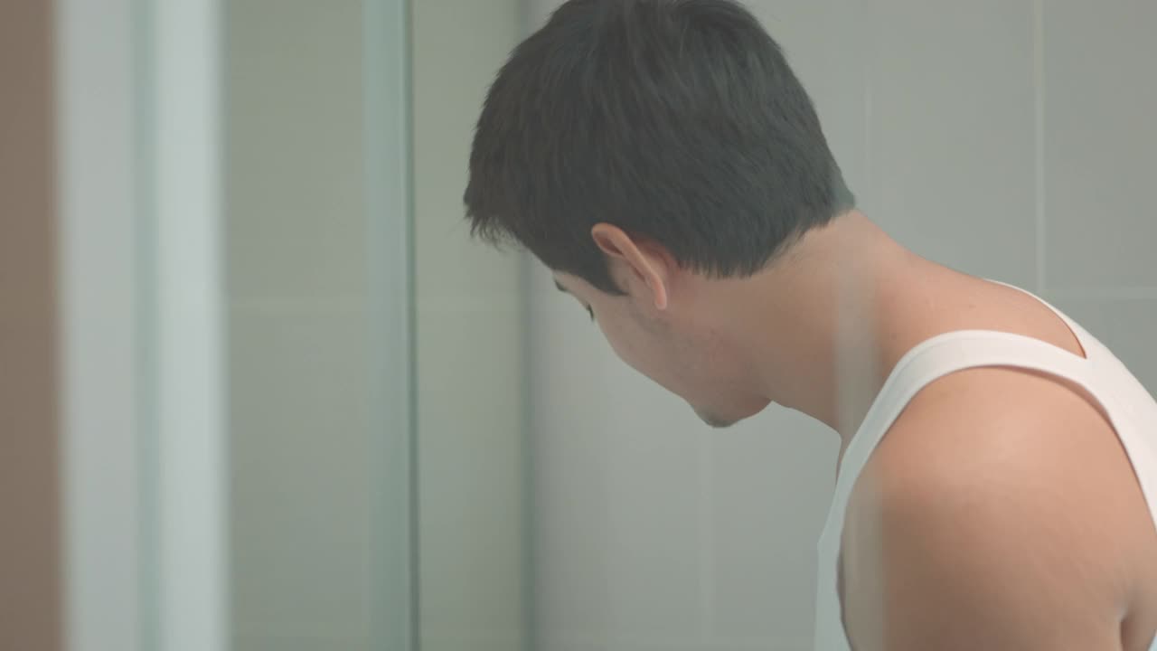 一名亚洲男子在洗澡后往脸上涂抹保湿面霜，准备在家里的浴室里工作。视频下载