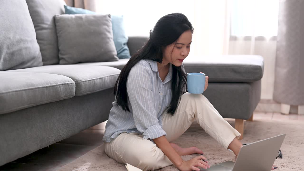 一位在客厅使用笔记本电脑的亚洲女性。商业女性在笔记本电脑屏幕上思考一个新项目，在家里工作的地方喝咖啡的肖像。在家工作，商业女性和工作理念。视频素材