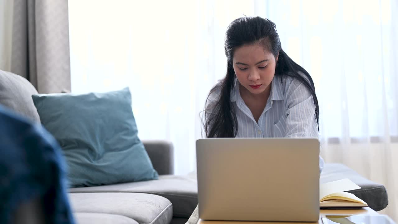 年轻的亚洲女商人正在笔记本电脑上寻找信息，她在笔记本上做笔记，她坐在家里客厅的沙发上。商业女性和工作理念。视频素材