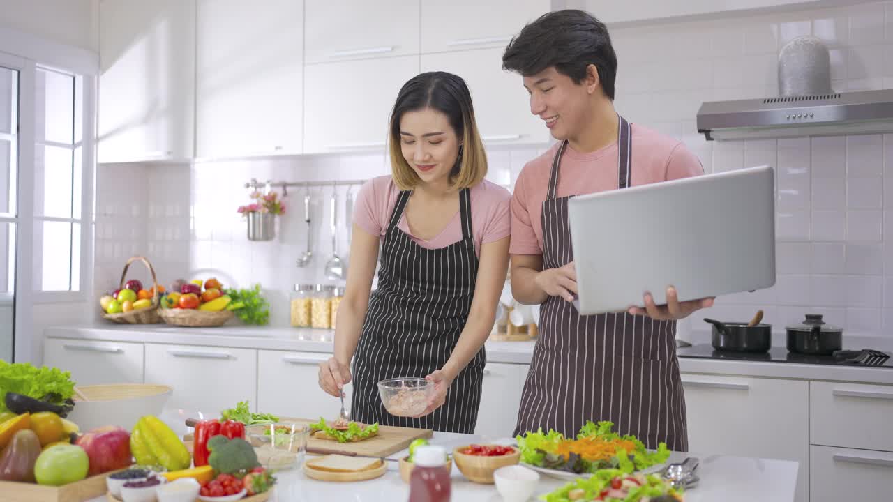 年轻的亚洲女子吃三明治做早餐烤面包在电烤箱。年轻人看着笔记本电脑和爱一起准备晚餐。健康食品和技术。视频素材
