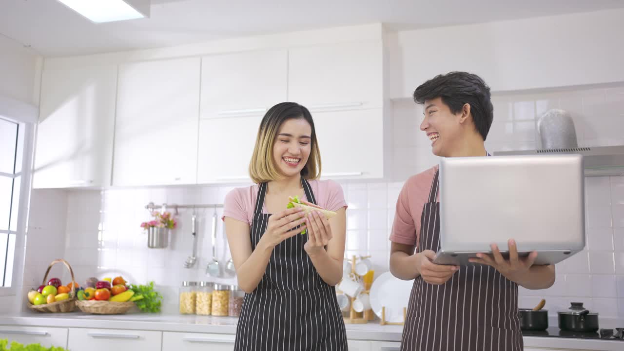 年轻的亚洲女子吃三明治做早餐烤面包在电烤箱。年轻人看着笔记本电脑和爱一起准备晚餐。健康食品和技术。视频素材