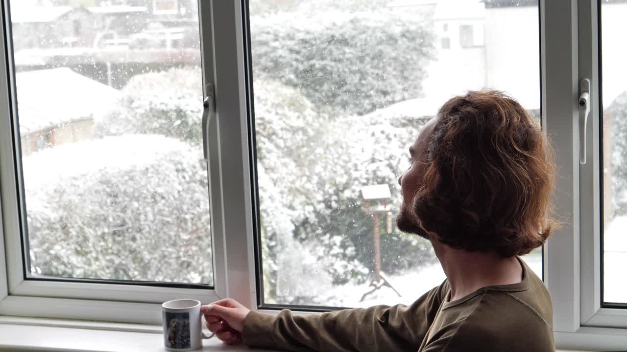 一个男人从窗框里看着雪花飘落，从杯子里喝水视频下载
