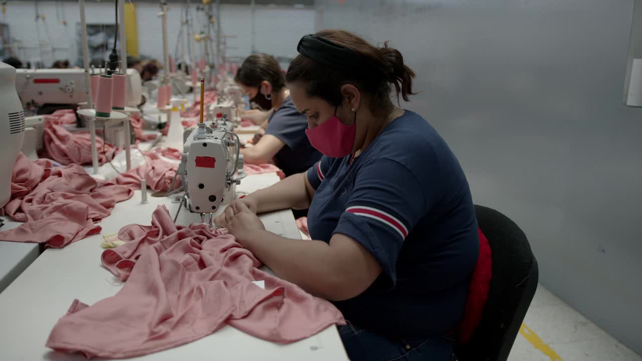 一群在服装厂的女工在缝纫站工作，她们都戴着防护口罩视频下载