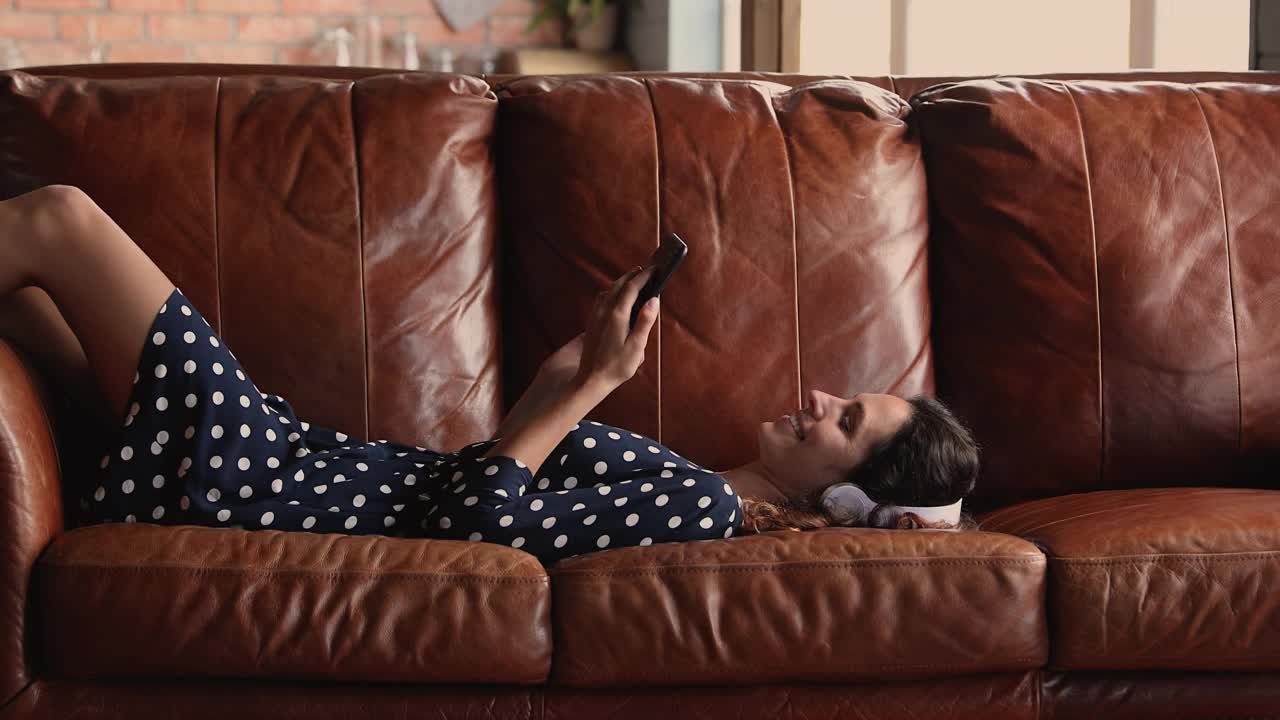 女人躺在沙发上戴着耳机用智能手机听音乐视频下载