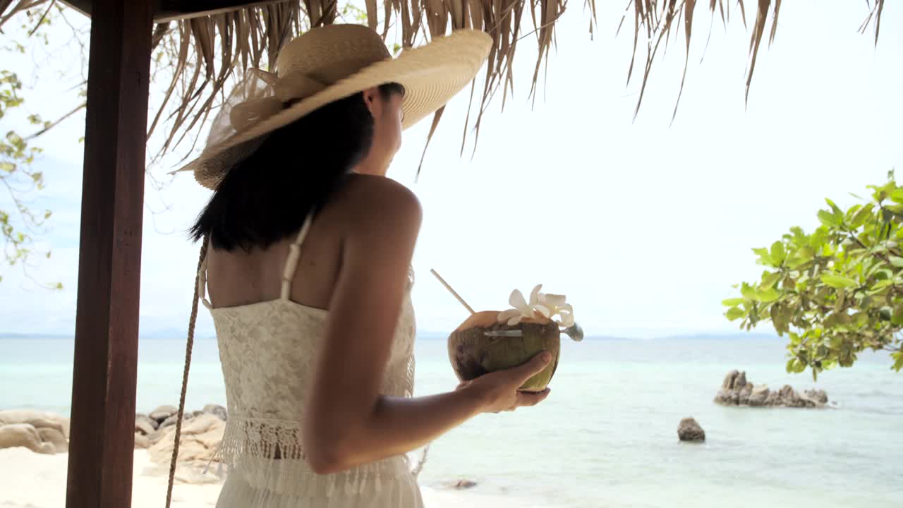 美丽年轻的亚洲女人喝椰子汁沿着海滩。视频下载
