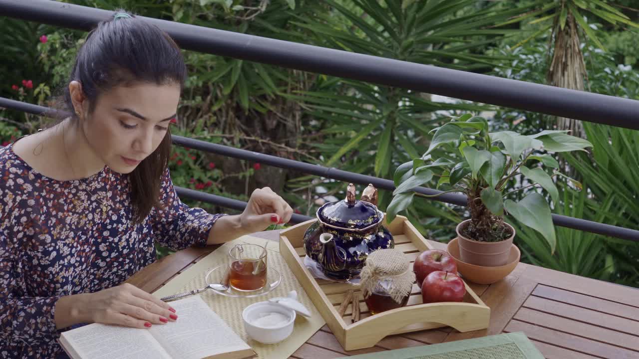 高角度的近景西班牙年轻女子喝一杯热茶，而阅读在户外露台的一本书视频素材