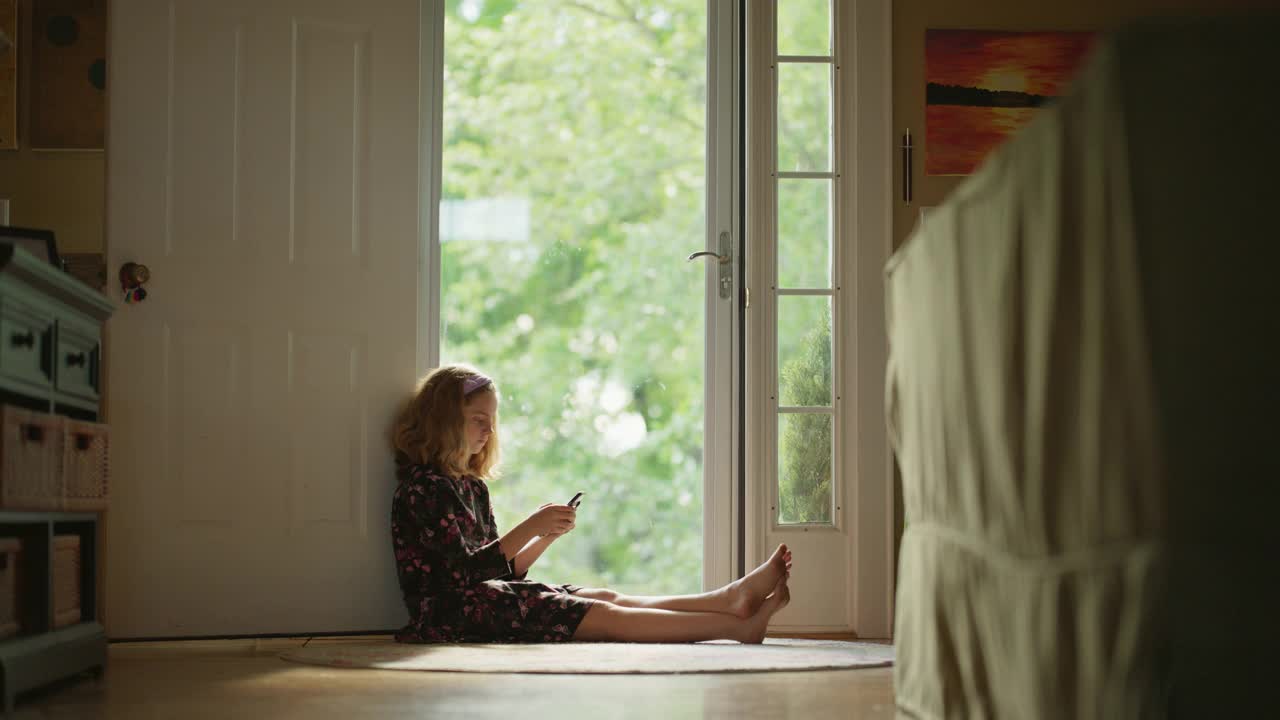一个年轻女孩坐在她的前门使用她的智能手机视频下载