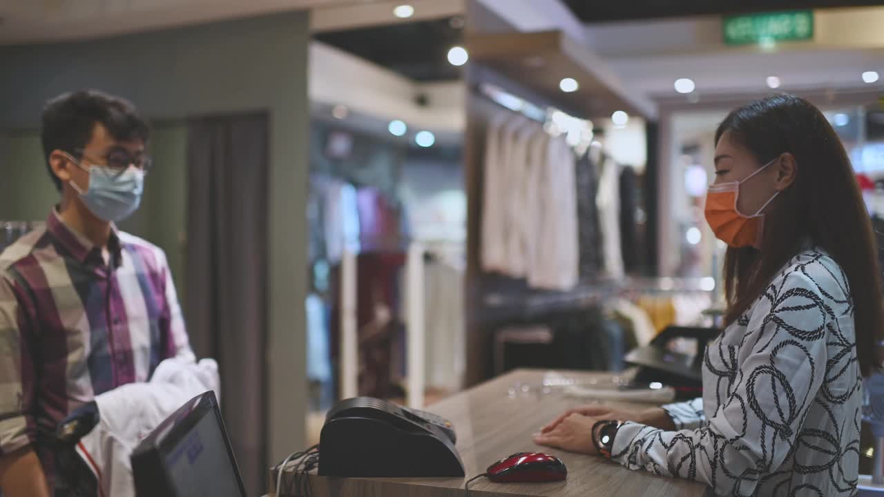 亚洲华人美丽的女男装店店主收银员用口罩折叠衣服，她的顾客把他们放在收银台的袋子里视频下载