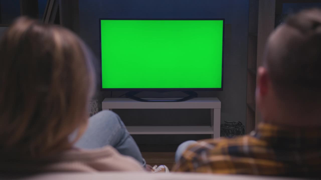 一对家庭夫妇一起坐在客厅的沙发上看绿屏电视模型。国内电影中看绿屏电视的休闲人士的后视观。在家休息看电视节目或新闻视频素材
