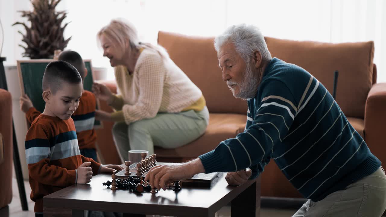 爷爷教孙子下棋，奶奶和孙子在家里的棋盘上画背景。有祖父母的双胞胎男孩，家庭娱乐的免费日视频素材