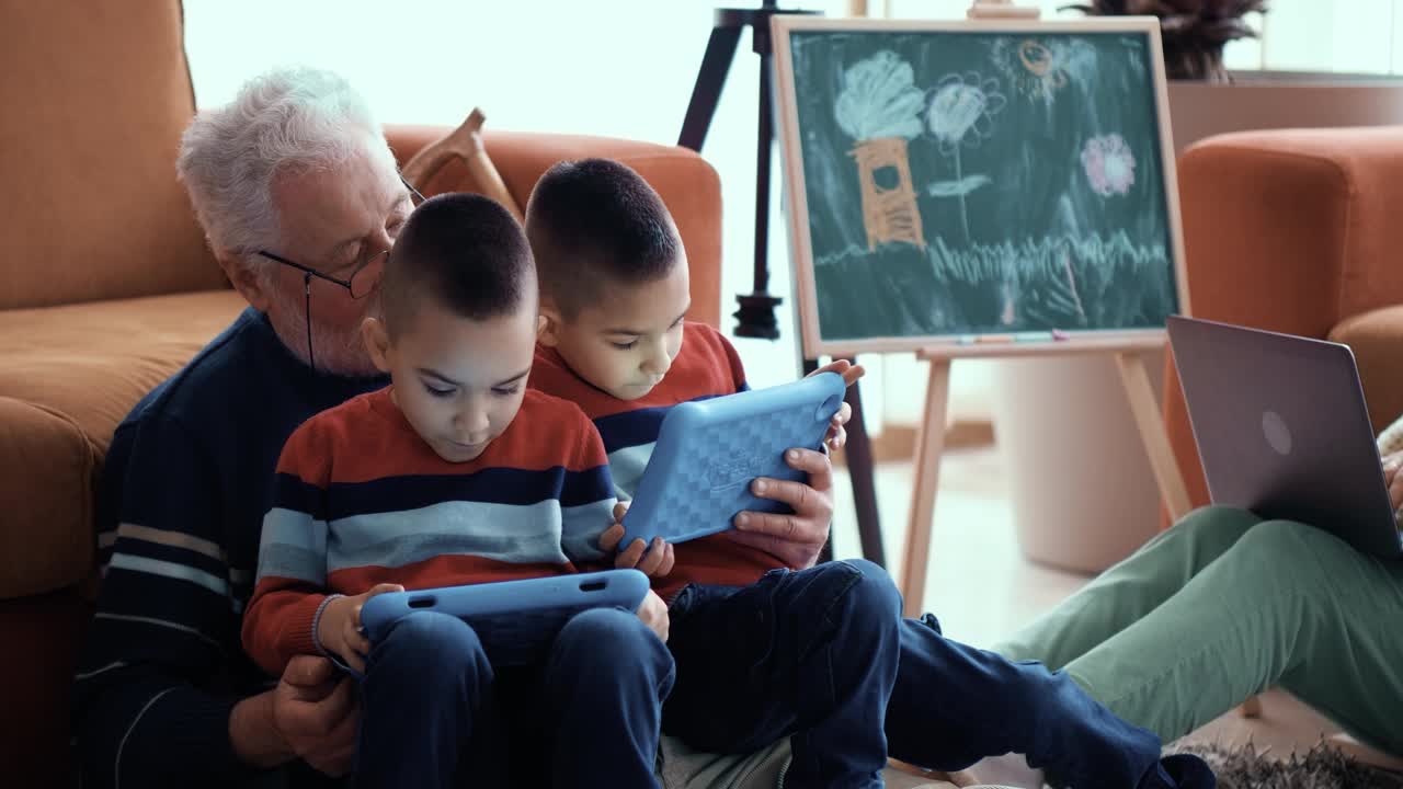 双胞胎孙子坐在爷爷的腿上玩平板电脑游戏，年轻的祖母用笔记本电脑，而爷爷娱乐小男孩视频素材