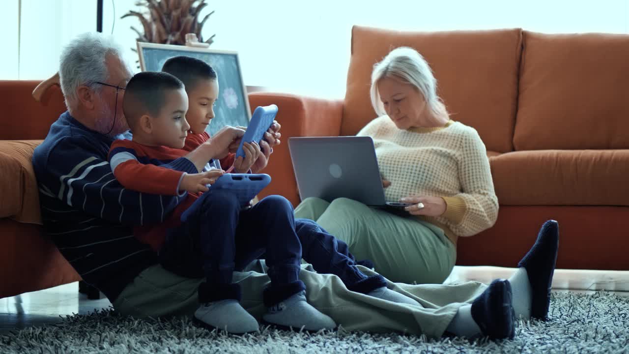 爷爷使用平板电脑，向孙子们展示如何玩游戏，奶奶在笔记本电脑上打字。这对双胞胎男孩用现代科技和他们的祖父玩得很开心视频素材