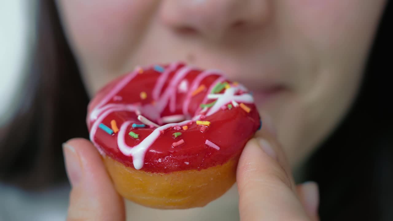 一个女人愉快地微笑着咬着一个软甜甜圈，上面覆盖着红色的水果釉和糖果屑。油腻的食物视频素材